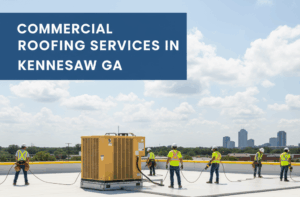 A team of roofers in hard hats and safety vests are working on a large flat commercial roof, installing roofing materials around a yellow HVAC unit. In the foreground, stacks of new roofing panels are visible. A city skyline can be seen in the distance under a partly cloudy sky.